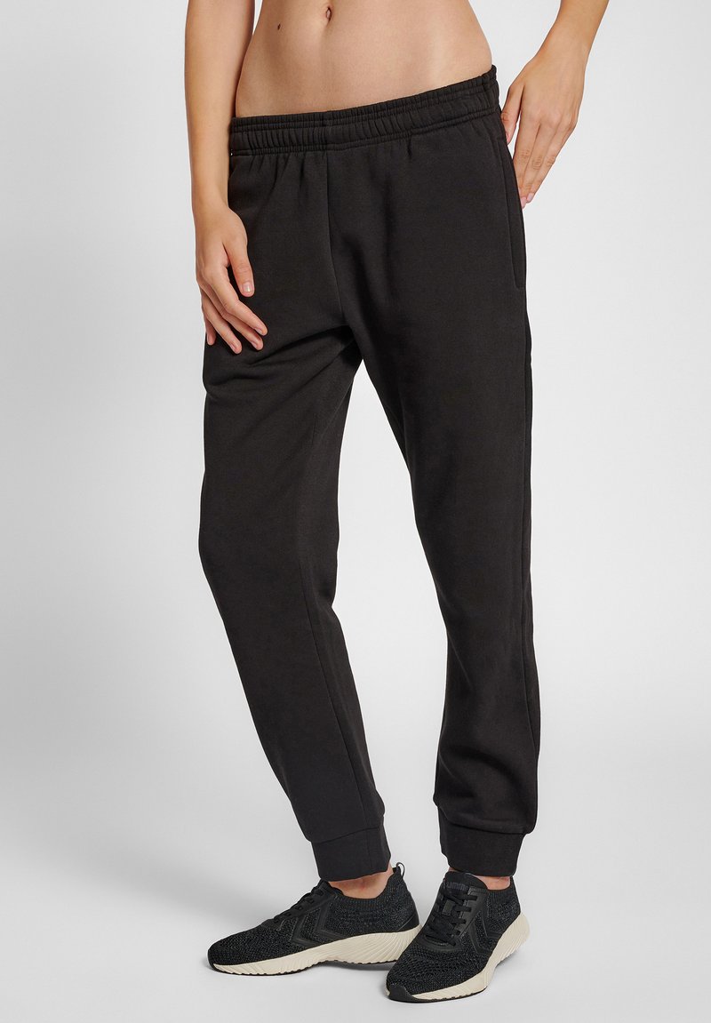 Strakke zwarte joggerbroek met elastische tailleband en omslag aan de enkels, gedragen met zwarte sportschoenen, getoond op een staand persoon.