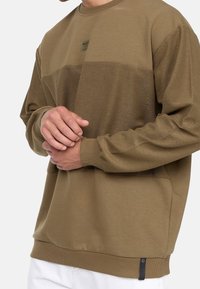 Brown sweatshirt with a textured horizontal stripe, ribbed cuffs and hem, featuring a small logo label on the bottom. Soft, fleece-like material.