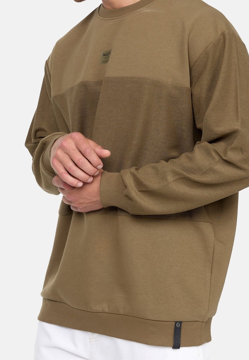 Brown sweatshirt with a textured horizontal stripe, ribbed cuffs and hem, featuring a small logo label on the bottom. Soft, fleece-like material.