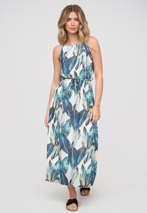 Femme portant une robe longue sans manches à imprimé feuilles tropicales bleues et vertes et des sandales noires, debout sur un fond uni.