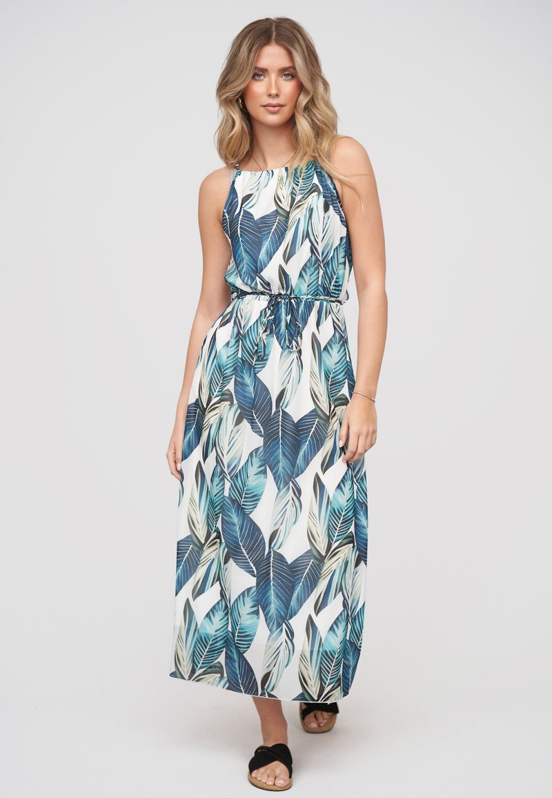 Femme portant une robe longue sans manches à imprimé feuilles tropicales bleues et vertes et des sandales noires, debout sur un fond uni.