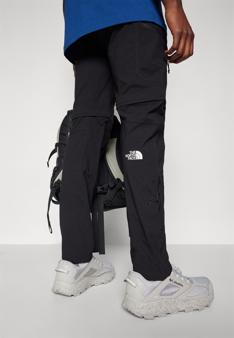 Czarne spodnie cargo z zamkami i kieszeniami, z logo "The North Face". Połączone z jasnoszarymi sneakersami i czarnym plecakiem.