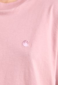 Hele roosa T-särk, millel on väike, ühevärviline broderi logo vasakul rinnaosal.