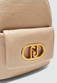 Primo piano di uno zaino in pelle beige con una superficie liscia, un pattina pieghevole e una chiusura con logo dorato in evidenza.