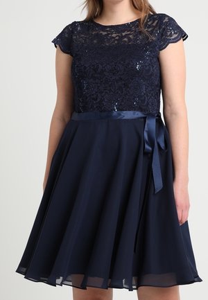 Vrouw draagt een marineblauwe jurk met kant en pailletten op het lijfje, een satijnen tailleband gebonden in een strik en een knielange vloeiende rok.