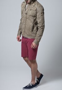 Kaki utilityjakke med flere lommer, parret med bordeaux shorts. Båret med marineblå sneakers, der har hvide snørebånd og gummisåler.