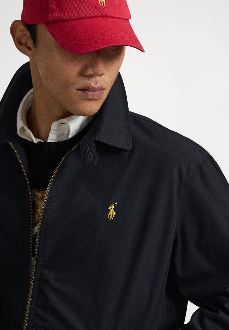Veste noire à fermeture éclair avec un logo polo doré, portée sur un pull foncé et une chemise blanche à col. Une casquette rouge complète le look.