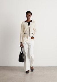 Cardigan lavorato a maglia crema con accenti neri, top nero, pantaloni bianchi a gamba larga e borsa nera, indossati con tacchi neri.
