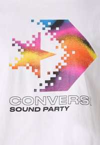 Camiseta blanca de algodón con un gráfico de estrella pixelada en varios colores que incluye tonos de azul, rosa, naranja y amarillo. El texto dice "CONVERSE SOUND PARTY."