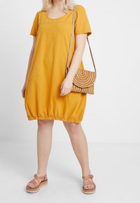 Kortärmad gul klänning med rundad halsringning och rynkad fåll, kombinerad med en vävd brun axelremsväska och ljusbruna sandaler.