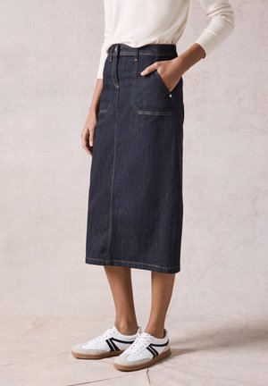 Personne portant une jupe midi en denim bleu foncé avec des poches frontales, un haut blanc à manches longues et des baskets blanches avec des rayures noires.