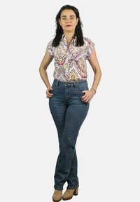 Blusa corta de manga corta con estampado floral en rosa y amarillo, combinada con unos ajustados vaqueros azules y botas de tacón color beige. Pose de pie.