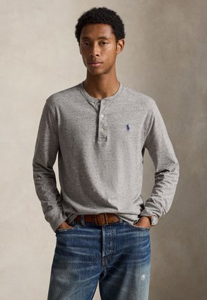 Homme portant un t-shirt henley gris à manches longues avec trois boutons, jean bleu et ceinture marron, debout les mains dans les poches.