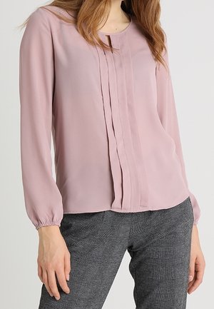 Vrouw draagt een stoffig roze bloes met lange mouwen en verticale plooien, gecombineerd met grijze geruite broek, tegen een effen achtergrond.
