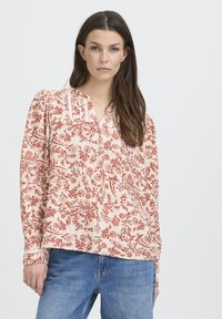 Femme aux longs cheveux bruns portant une blouse légère à motifs floraux rouges et un jean bleu, se tenant devant un fond blanc uni.