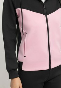 Veste à capuche zippée présentant un panneau supérieur noir et un panneau inférieur rose, fabriquée en tissu lisse avec des poches avant et des cordons de serrage ajustables.