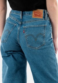 Jean en denim bleu avec une coupe droite, comportant une étiquette de marque en cuir et une couture courbée distinctive sur les poches arrière.