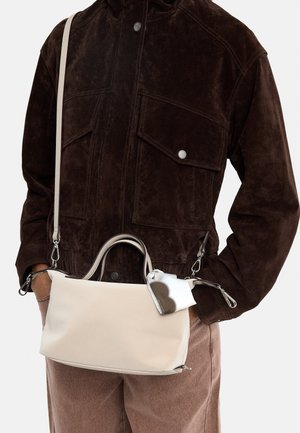 Osoba ubrana w ciemnobrązową zamszową kurtkę i jasnobrązowe spodnie, niosąca beżową torebkę z rączkami i dołączonym wzorzystym materiałem.