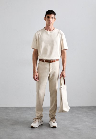 Tricou bej cu guler rotund, pantaloni bej deschis croiți, curea maro și adidași albi cu accente bej. Modelul poartă o geantă de umăr bej.