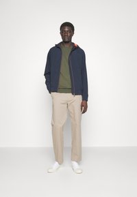 Jachetă bleumarin cu fermoar și accente portocalii, purtată peste un pulover verde, pantaloni bej și adidași albi. Design simplist cu o buzunar frontal.