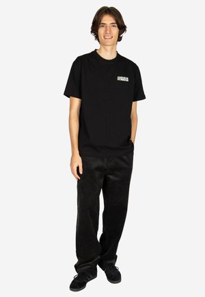 Young person standing, smiling, wearing a black t-shirt with a small logo and black corduroy pants against a plain light background.