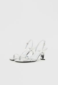 Ασημί μεταλλικά σανδάλια ανοιχτού δαχτύλου με λεπτές λωρίδες και κοντό κωνικό τακούνι, με γυαλιστερή φινίρισμα και ρυθμιζόμενο λουράκι στον αστράγαλο.