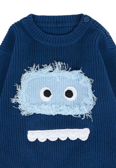 Pull en tricot bleu avec un patch duveteux bleu clair portant des yeux et un détail festonné blanc en bas. Col côtelé et boutons.