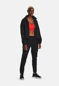 Giacca leggera nera, top sportivo rosso a maniche corte, pantaloni da jogging neri con polsini elasticizzati e scarpe da ginnastica nere con dettagli bianchi.