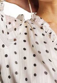 Blusa blanca con lunares negros, con un cuello rizado, rayas verticales y un ligero brillo. La tela parece ligera y texturada.