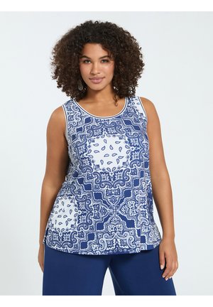 Femme aux cheveux bouclés portant un haut sans manches bleu et blanc à motifs et un pantalon bleu uni, debout devant un fond clair uni.