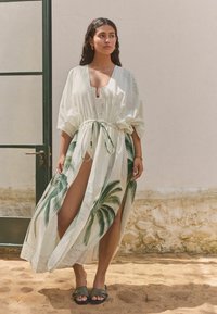 White kimono-style cover-up with palm tree print, tie waist, short sleeves, and thigh-high slit, worn over a light-colored dress and sandals.