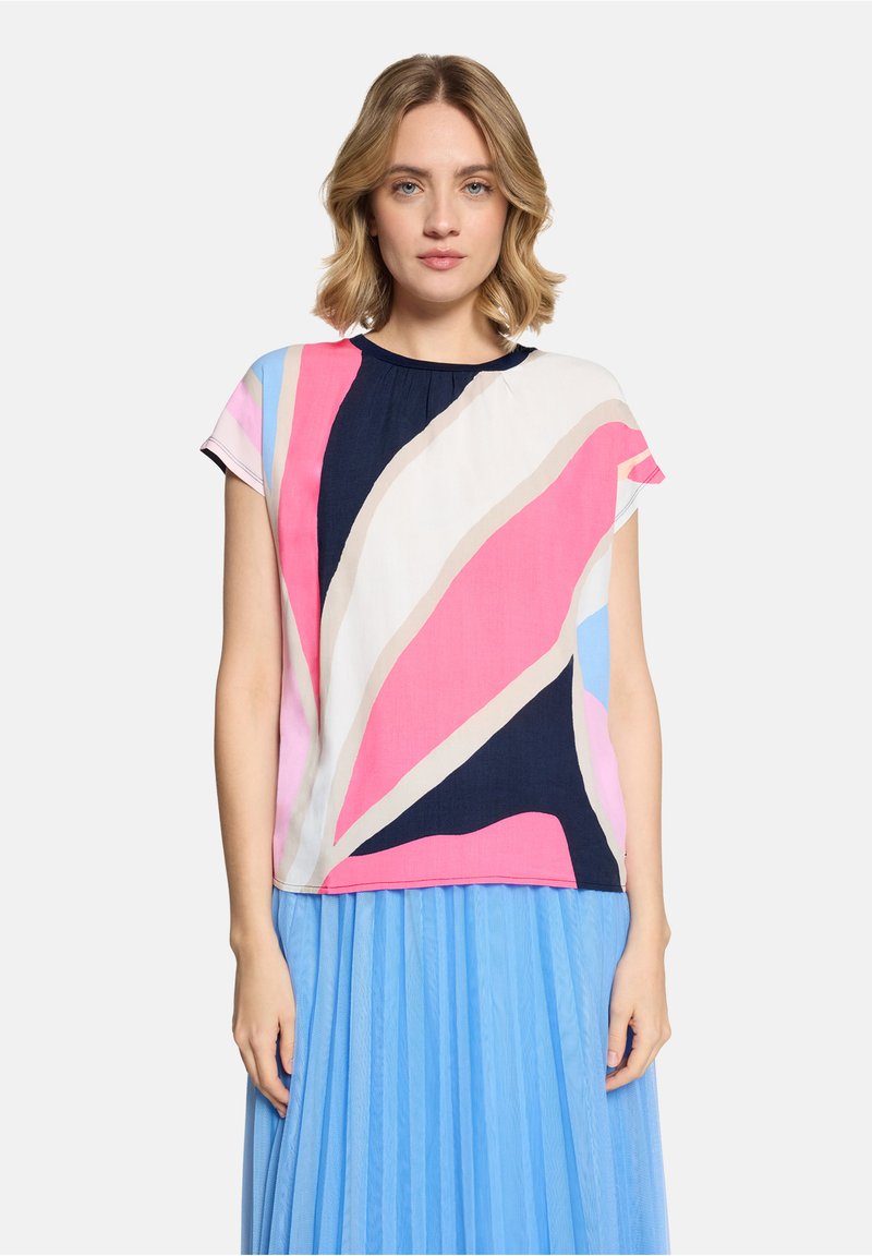 Femme aux cheveux blonds mi-longs portant un haut à motif abstrait multicolore et une jupe plissée bleu clair, debout devant un fond blanc.