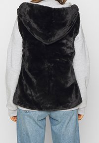 Czarna kamizelka z sztucznego futra z kapturem, charakteryzująca się miękką teksturą i luźnym krojem, noszona na szarym swetrze z długimi rękawami oraz na codziennych dżinsach.