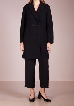 Vrouw met een zwarte dubbelgeknoopte jas, zwarte cropped broek en zwarte lage hakken, staand tegen een effen achtergrond.