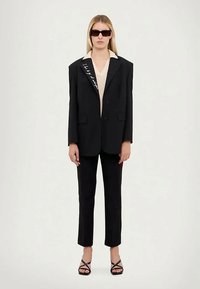 Woman wearing oversized black blazer with white script, black trousers, beige top, black sunglasses, and black strappy heels, standing against plain background.