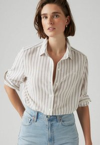 Witte blouse met verticale multicolore strepen, korte opgerolde mouwen en een kraag. Draagt het met lichtblauwe spijkerbroek.