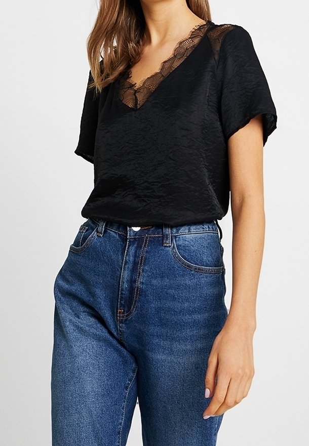 Femme portant un chemisier noir à manches courtes avec un col en V en dentelle et un jean bleu taille haute sur un fond uni.
