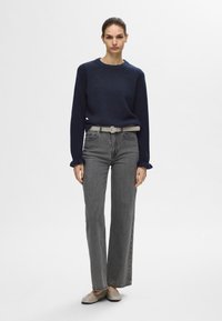 Mornarsko modra pletena majica z okroglim izrezom, sive kavbojke z visokim pasom in bež balerinke. Kavbojke so s krojem z ravnimi nogavicami in subtilnim bledenjem.