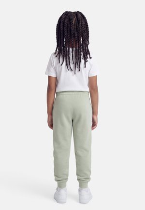 Copil cu păr lung și răsucit, purtând un tricou alb, pantaloni de trening verde deschis și adidași albi, stând cu fața spre spate pe un fundal simplu.