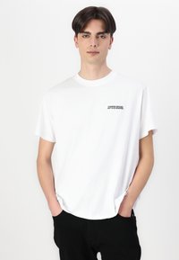 Camiseta de algodón blanca con cuello redondo y mangas cortas, con un logo impreso en negro en el lado izquierdo del pecho. Se lleva con pantalones negros.