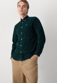 Camisa de cotê verde-escuro com colarinho abotoado, apresentando um bolso no peito e um padrão xadrez subtil, combinada com calças beje claras.
