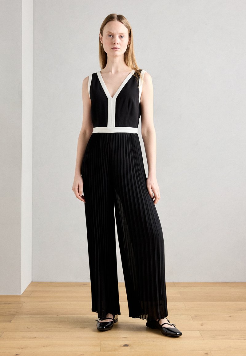 Jumpsuit noir plissé avec un décolleté en V et une taille blanche. Présente des bretelles fines et une silhouette fluide. Porté avec des chaussures plates noires.