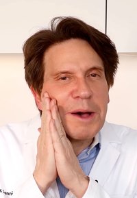 Medisinsk fagperson i hvit frakk, med mørkt hår, sammenflettede hender foran ansiktet. Enkel lys bakgrunn, fokusert ansiktsuttrykk.