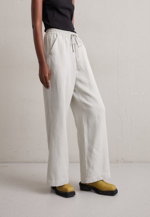 Personne portant un pantalon blanc ample à cordon, un haut noir sans manches et des chaussures compensées jaune moutarde, debout devant un mur clair uni.