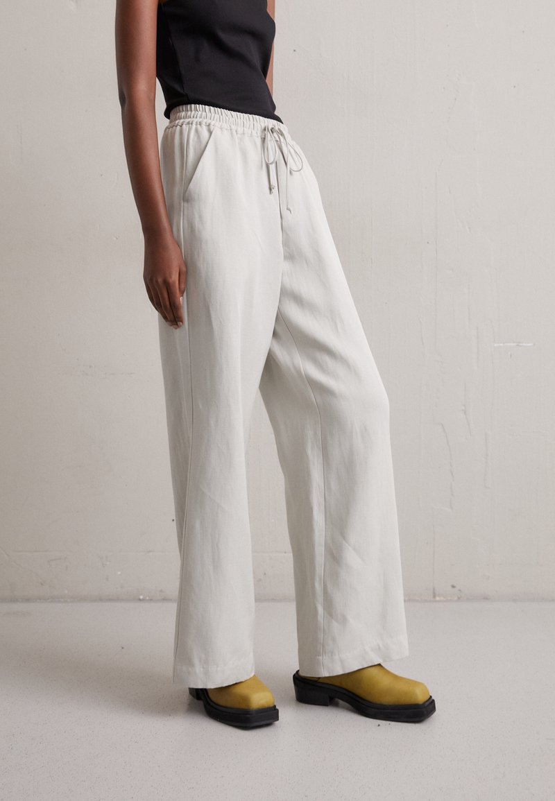 Personne portant un pantalon blanc ample à cordon, un haut noir sans manches et des chaussures compensées jaune moutarde, debout devant un mur clair uni.