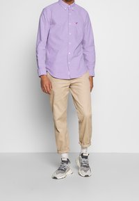 Camicia button-up viola chiaro con taschino e logo ricamato, abbinata a pantaloni beige a risvolto e sneakers grigie. Un outfit semplice e informale.