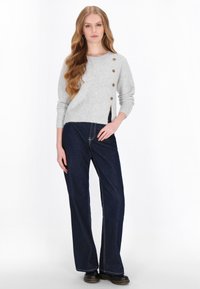 Lys grå striktrøje med en sideslids og fire brune knapper, parret med mørkeblå brede jeans med kontrastsyninger.