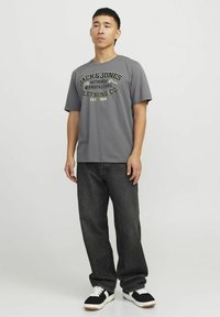 Grijze korte mouwen T-shirt met zwart en gele 'JACK & JONES' grafische print, gecombineerd met donkerdenim jeans en zwarte sneakers met witte accenten.