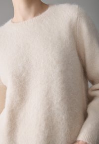 Lys beige genser laget av fluffy materiale med rund hals og lange ermer, med en myk, teksturert overflate.