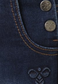 Jeans in denim blu scuro con cuciture arancioni a contrasto, bottoni in metallo e un sottile logo di ape ricamato sulla tasca.